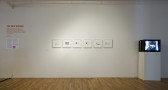A gallery wall with six framed images of passport copies. On the right side of the images, a television stands on a white plinth, showing a black and white image. On the left side, wall text lists the exhibition title and artists.