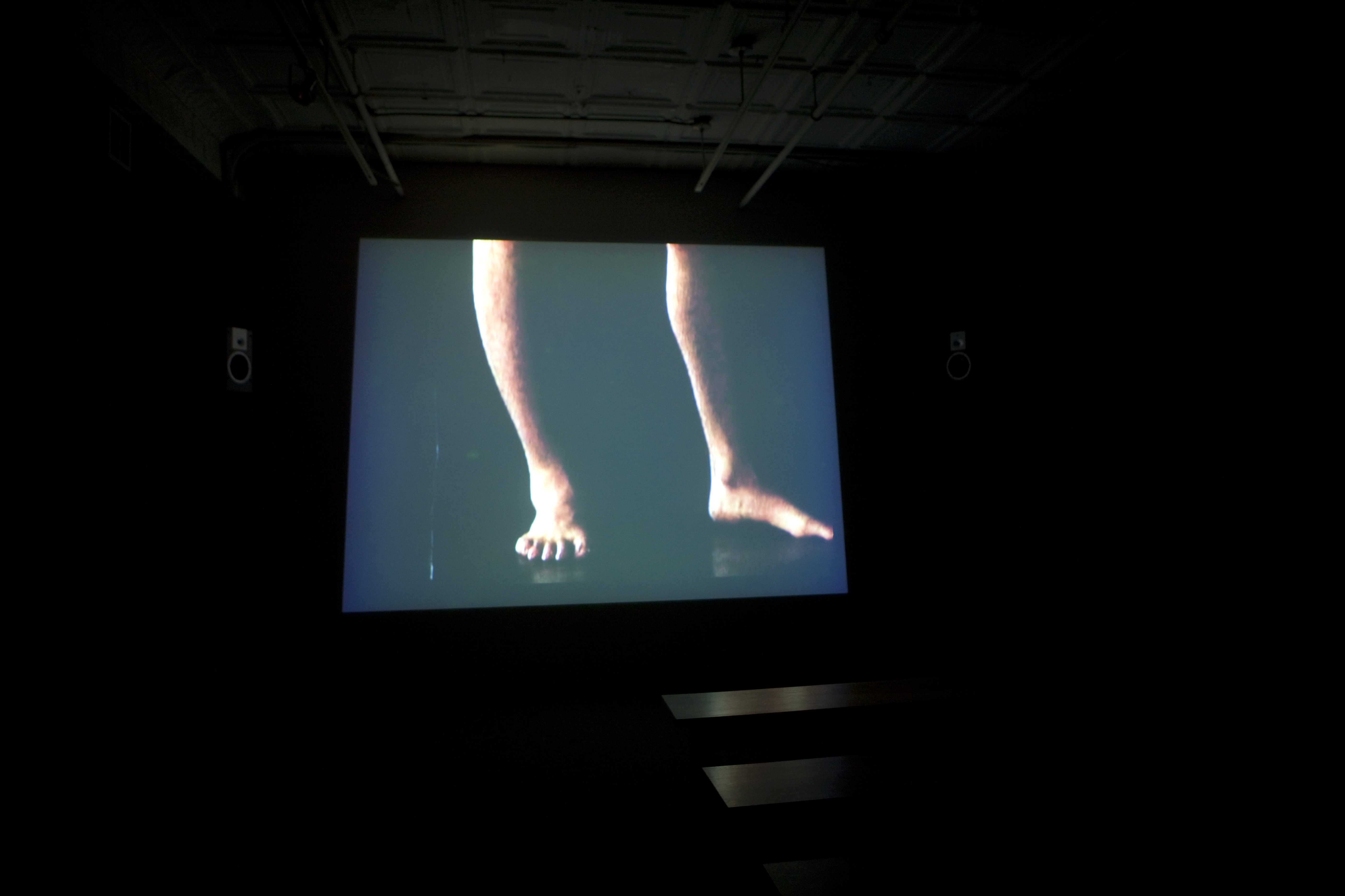 A large screen in a dark room, showing an image of a pair of bare legs from the knees down.
