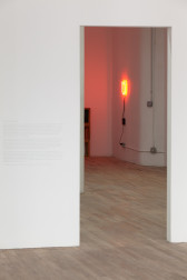 A red, glowing neon light installed on a gallery wall just above eye-level is visible through a doorway opening in a white-walled space. To the left of the opening is a block of faint text on the gallery wall.