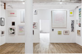 Several framed posters, photographs, and other ephemera of varying sizes hang on a white gallery wall. At the center of the wall is a doorway-shaped opening, through which more of the gallery space is visible.