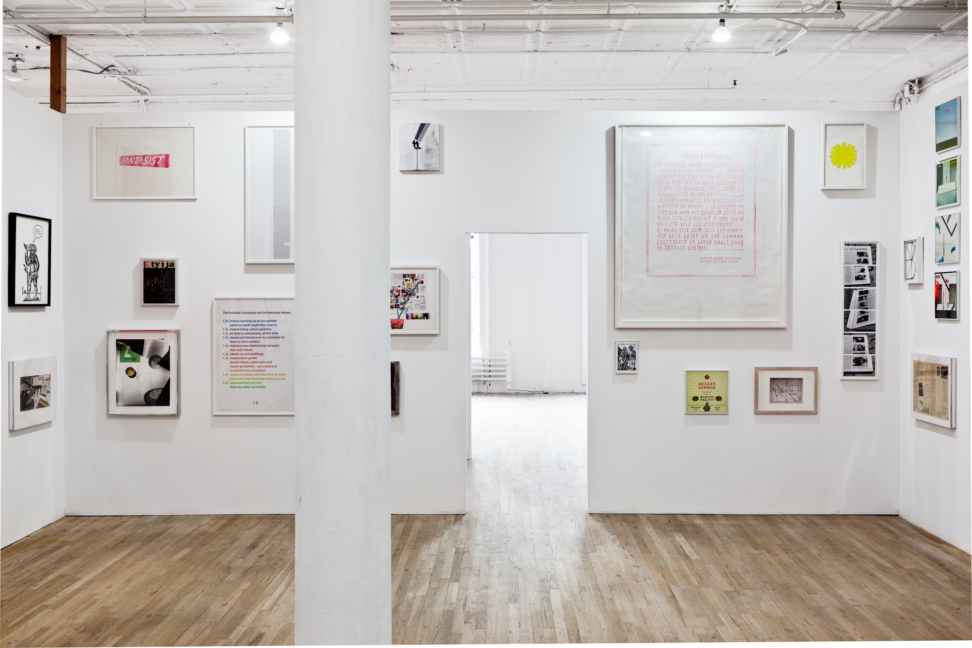 Several framed posters, photographs, and other ephemera of varying sizes hang on a white gallery wall. At the center of the wall is a doorway-shaped opening, through which more of the gallery space is visible.