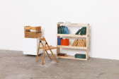 In an otherwise empty, white-walled room a wooden folding chair faces a wooden bookshelf with three shelves. Several clusters of colorful books rest on the bookshelf. To the left of the bookshelf is a square white bin which holds several more folding chairs.