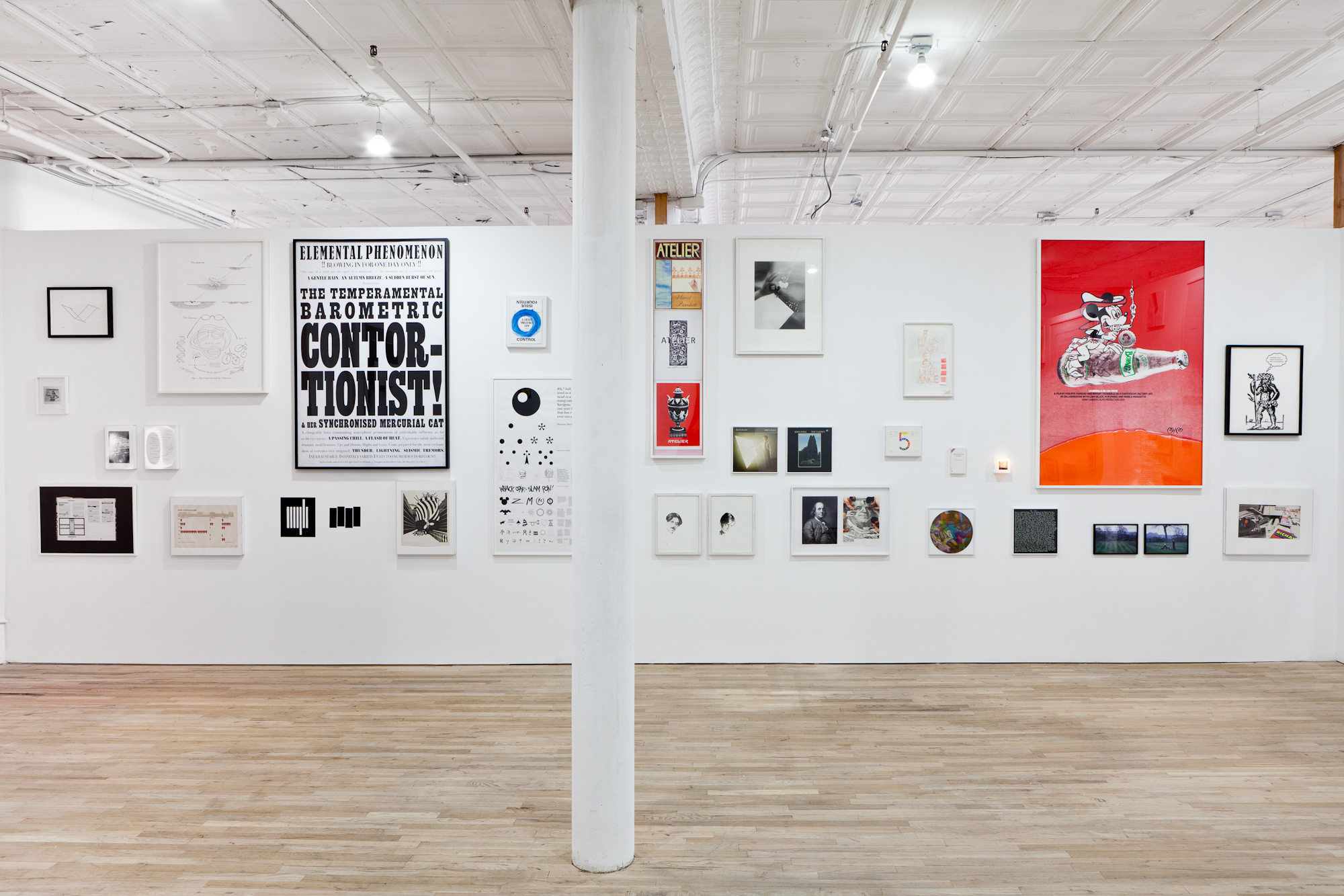 A large number of framed posters, photographs, and other ephemera of varying sizes hang on a white gallery wall. A poster hung on the left side of the wall has large black lettering that reads: 