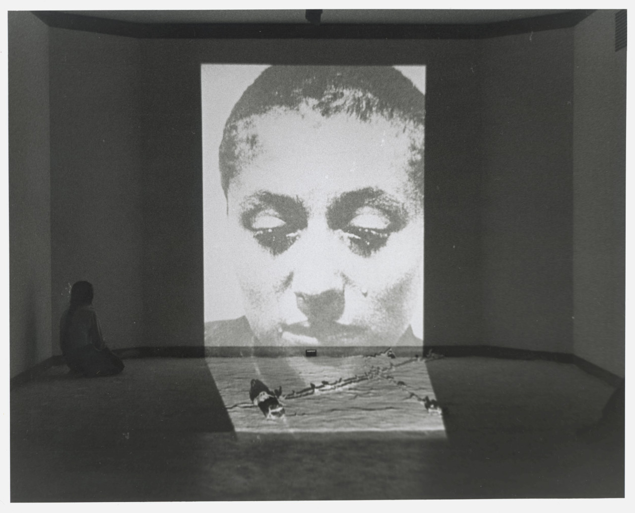 Black-and-white photograph of a wall with a large projection of a woman's face on it. To the left of the projection, a woman with dark hair kneels on the floor facing the projection.