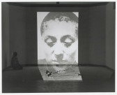 Black-and-white photograph of a wall with a large projection of a woman's face on it. To the left of the projection, a woman with dark hair kneels on the floor facing the projection.
