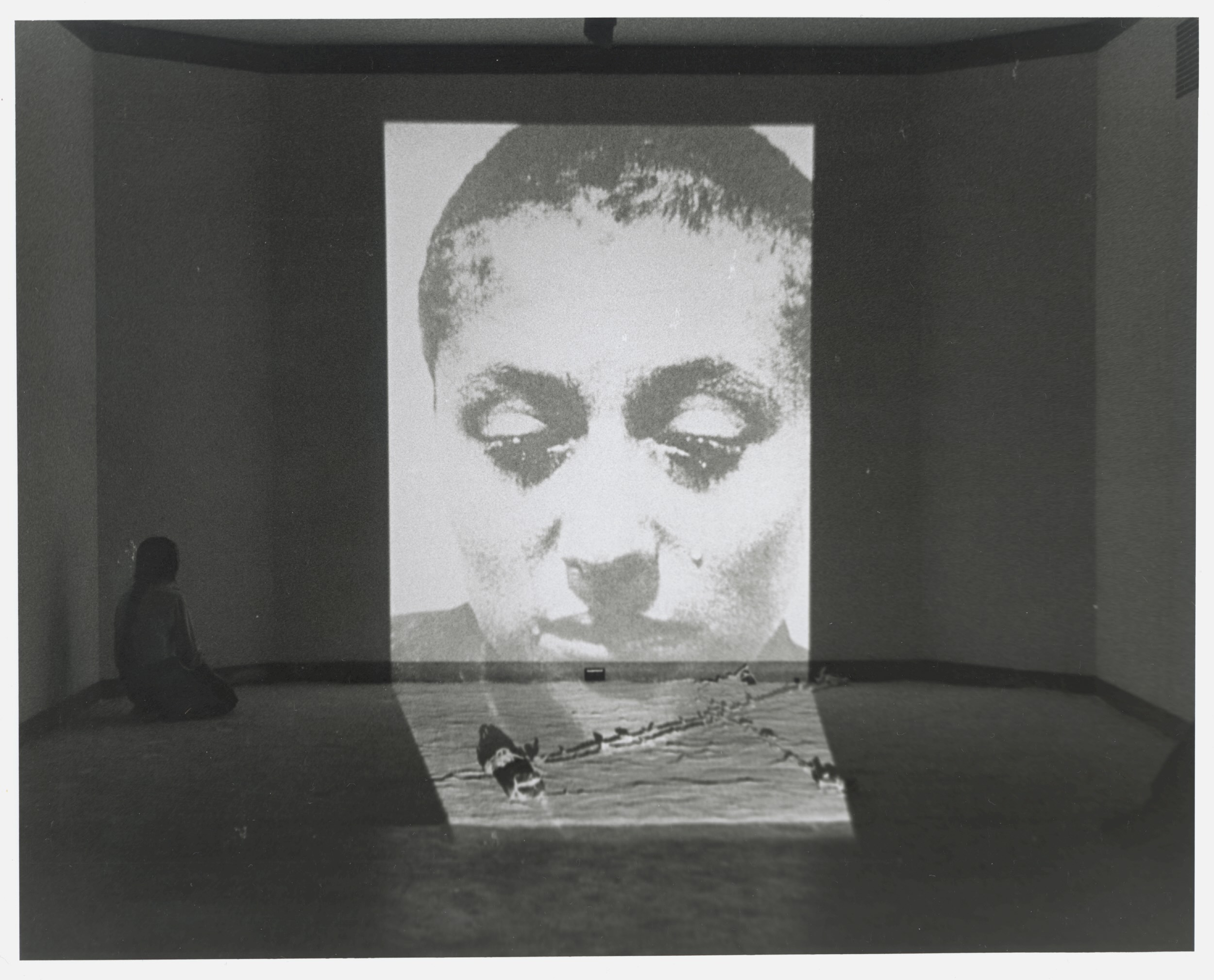 Black-and-white photograph of a wall with a large projection of a woman's face on it. To the left of the projection, a woman with dark hair kneels on the floor facing the projection.