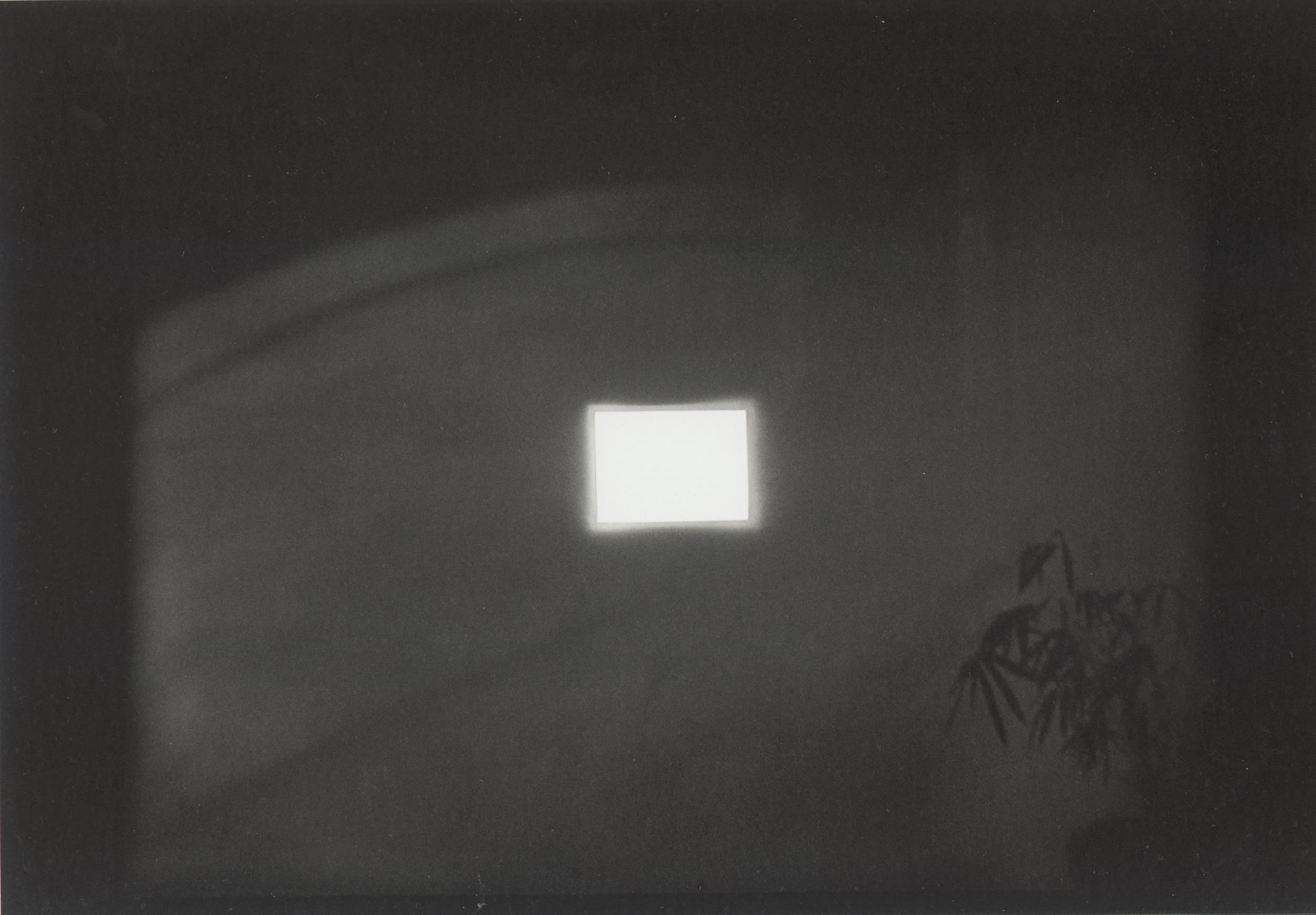 Black-and-white photograph of a projected image of shadows against a wall. In the center of the wall a bright light shines through a rectangular cut-out.