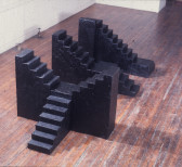 A color installation of three black tiered sculptures, placed on a wooden floor in a gallery space.