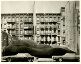A black and white film still of an apartment building, taken from the street. In front of the building is a sculpted landscape of rolling green grass. Two cars are parked on the street.