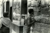A black and white film still of two figures on a sidewalk, one stands against a stone wall looking through a small opening in the stone.