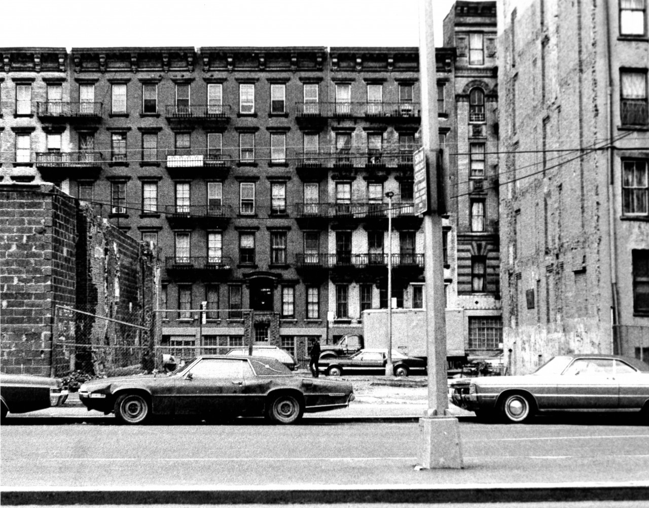 A black and white film still of an apartment building, taken from the street. In front of the building is an empty lot. Two cars are parked on the street.