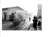 A city street is visible with a items and people driving by and a motorcyclist. On the sidewalk are people walking toward the camera, but looking in different directions. On the building behind the road is writing that reads, 