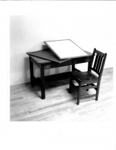 An image of a wooden chair next to a wooden desk with a frame being prepared on top of it. They are placed on a wooden floor against a white wall.