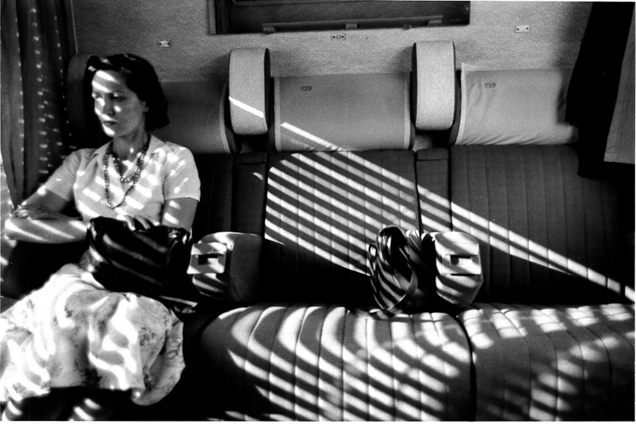 A figure sits nexts to a window with light shining through the shades, making an evident striped shadown on them and the seats next to them, one of which includes a purse.