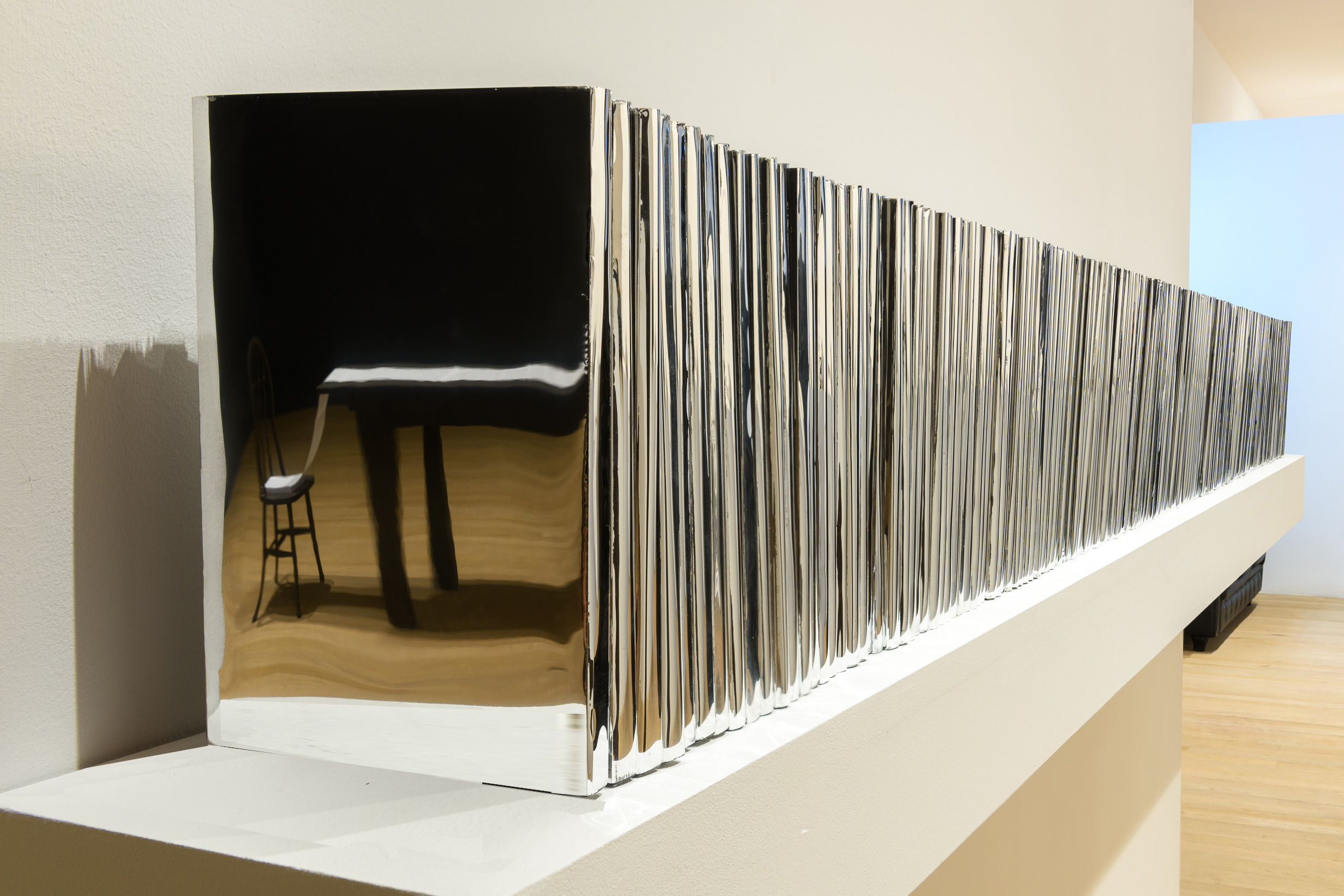 A row of books with silver covers on a shelf. A black table and chair can be seen in the reflection of the book cover.