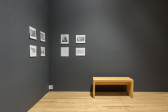 A photograph of a gallery space with dark grey walls. Eight photographs and print-outs are hung on the walls, and to the right of them is a wooden bench.