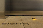 A close-up photograph of an orange LED light on the gallery floor. Black vinyl on the floor in front of the LED reads, 