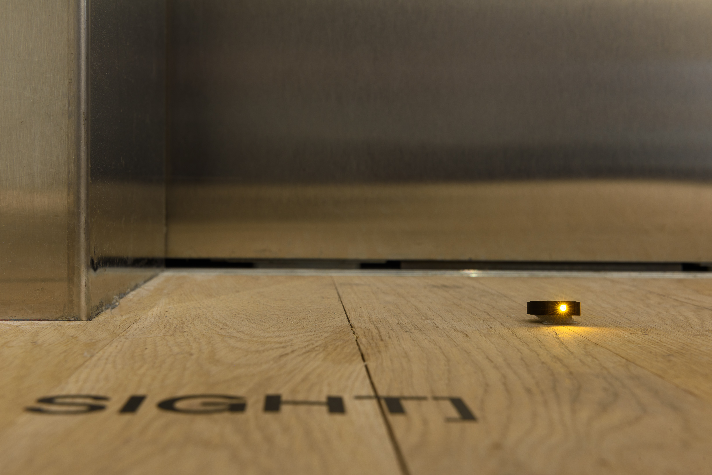 A close-up photograph of an orange LED light on the gallery floor. Black vinyl on the floor in front of the LED reads, 