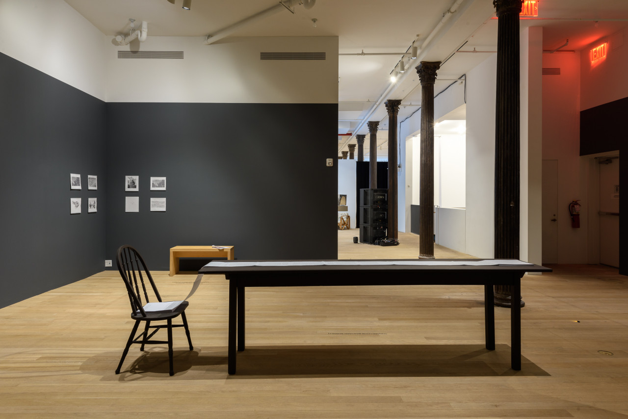 A photograph of a gallery space with a black rectangular table and a chair in the foreground. There are eight photographs and print-outs hung on the back walls, and a row of floor-to-ceiling columns is visible in the background.