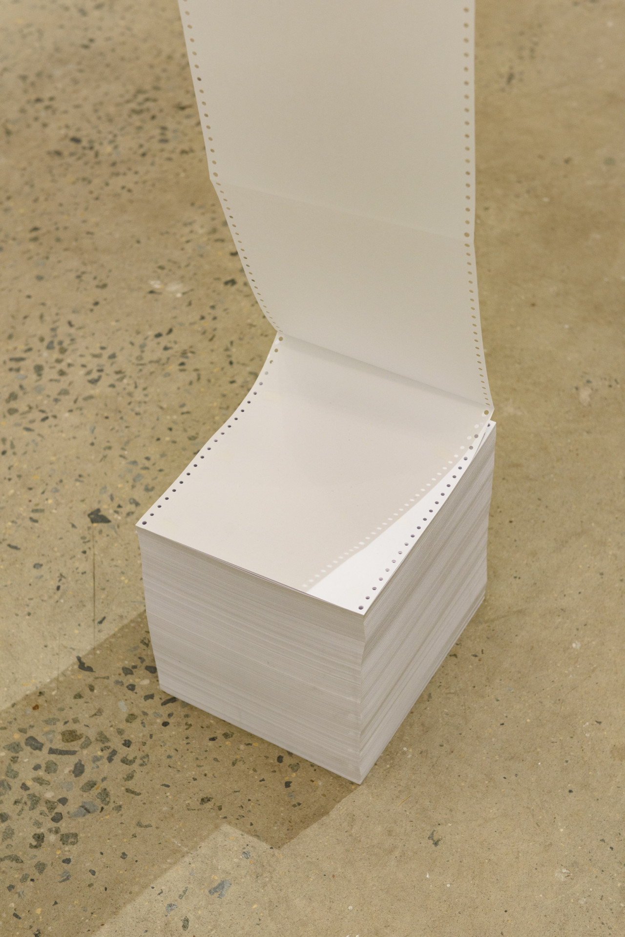 A long sheet of white paper, hung vertically, is attached to a stack of paper on the gallery floor.