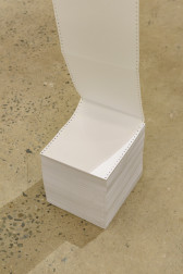 A long sheet of white paper, hung vertically, is attached to a stack of paper on the gallery floor.