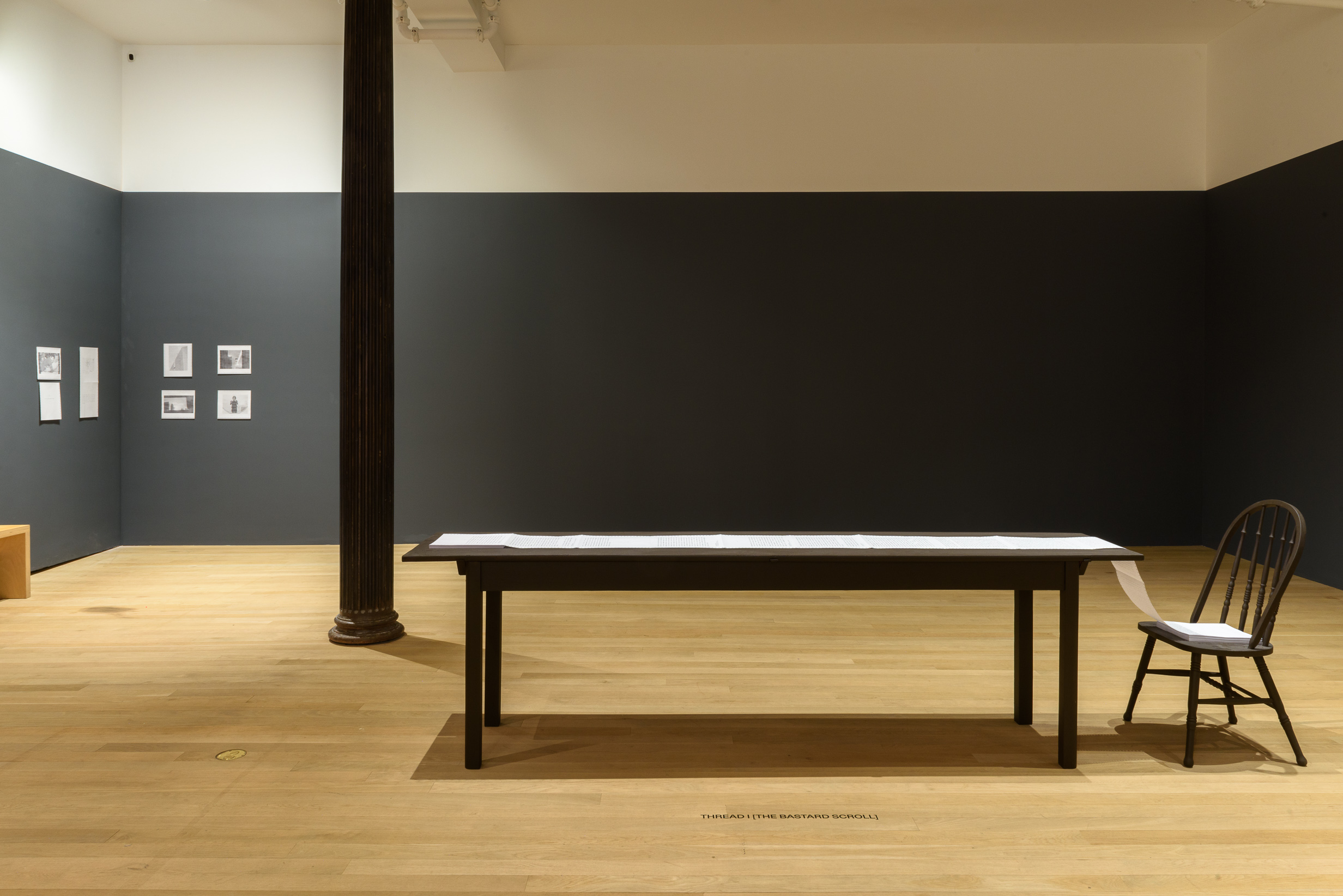 A photograph of a gallery space with a long rectangular table and a black chair. On the table is a long sheet of white paper. In the corner hangs eight photographs and print-outs next to a wooden bench.