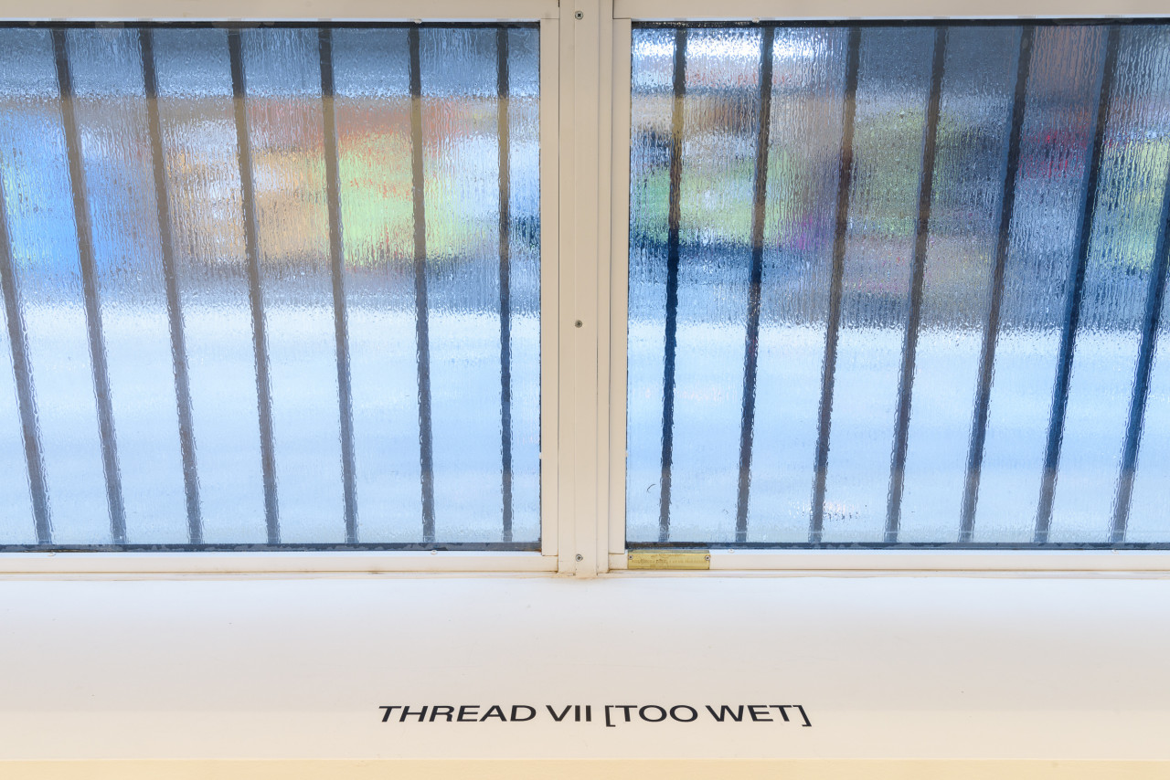 A photograph of a foggy gallery window. The words on the windowsill read "THREAD VII (TOO WET)."