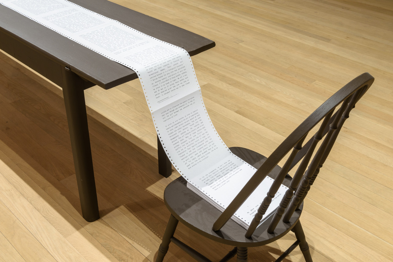A photograph of a black table next to a black chair. The end of a long sheet of white paper on the table rests on the chair.