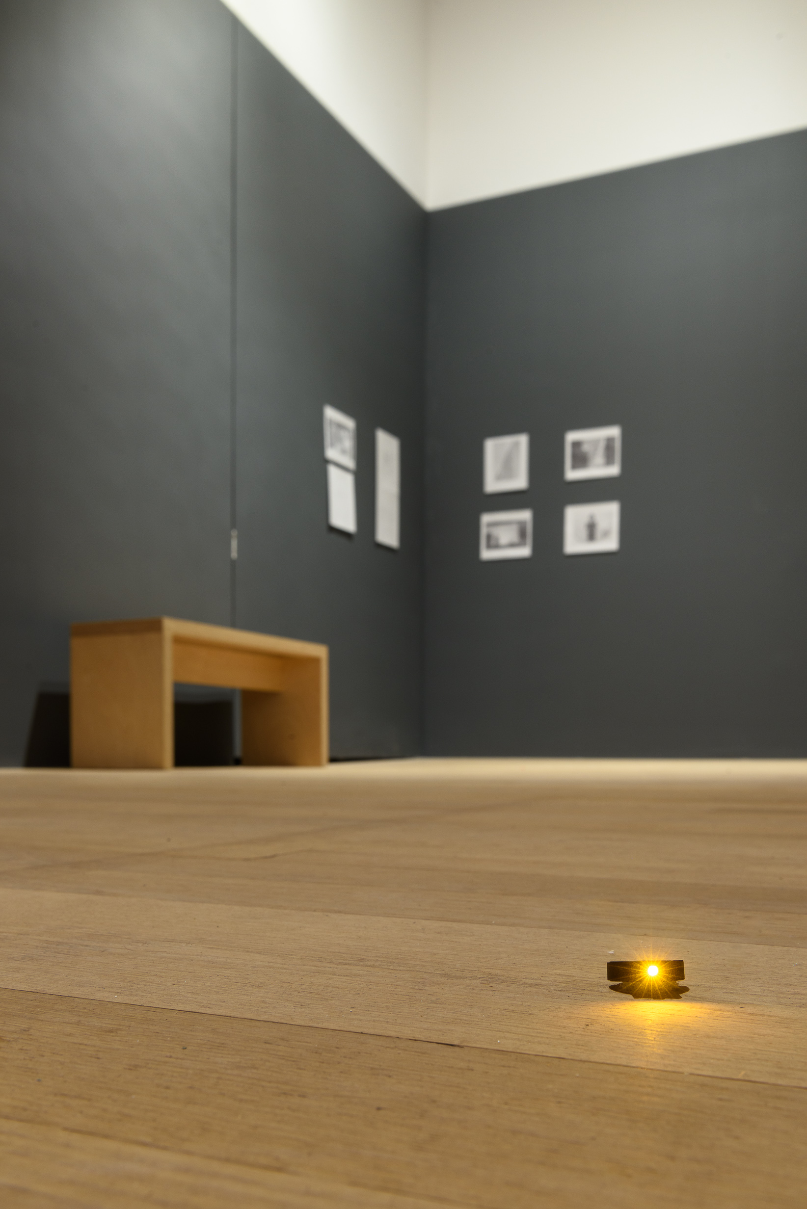 A photograph of a gallery space with eight photographs and print-outs hung on the walls next to a wooden bench. In the foreground, there is an orange LED light on the floor.