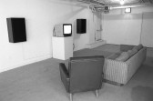 A black and white image of two couches set in front of a TV monitor on a pedestal and two speakers installed on either side of it. The TV screen is on, but no clear image is visible on it. A door to exit is visible in the background.