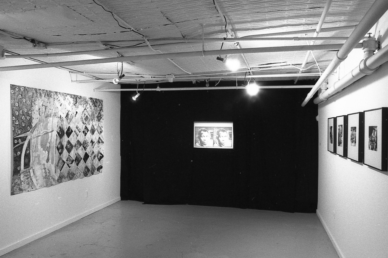 A black and white photograph of a mixed media collage hanging on a gallery wall hanging opposite of four mixed media collaged in frames with a back-projected slide image of two faces centerd in the image with a dark curtain covering its surroundings.