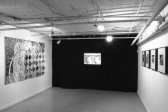 A black and white photograph of a mixed media collage hanging on a gallery wall hanging opposite of four mixed media collaged in frames with a back-projected slide image of two faces centerd in the image with a dark curtain covering its surroundings.