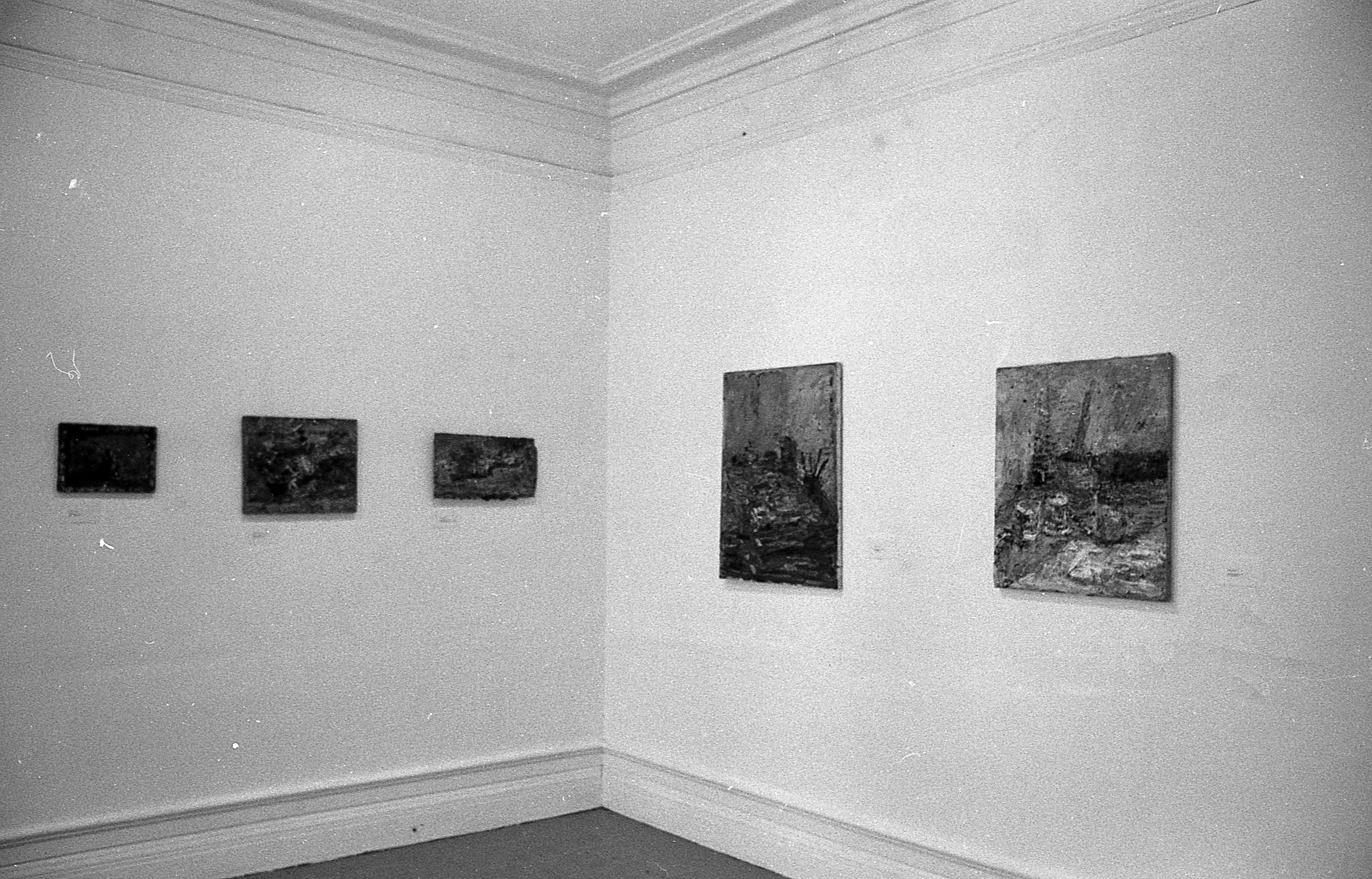 5 paintings hang in a corner with 3 on the wall to the left and 2 on the wall to the right.