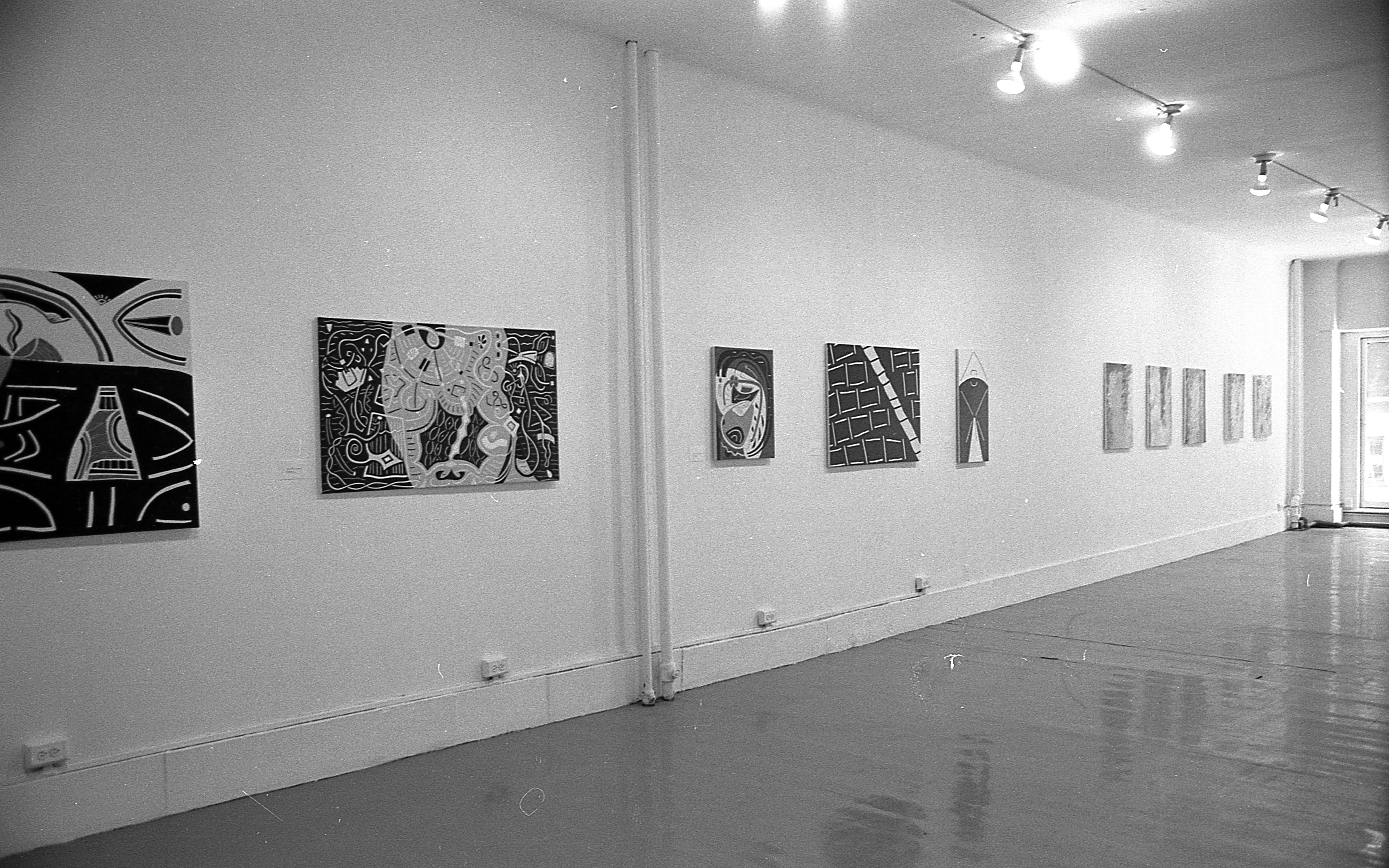 Various paintings hang in a row on a wall. The paintings are composed of bold lines and blocks of color.