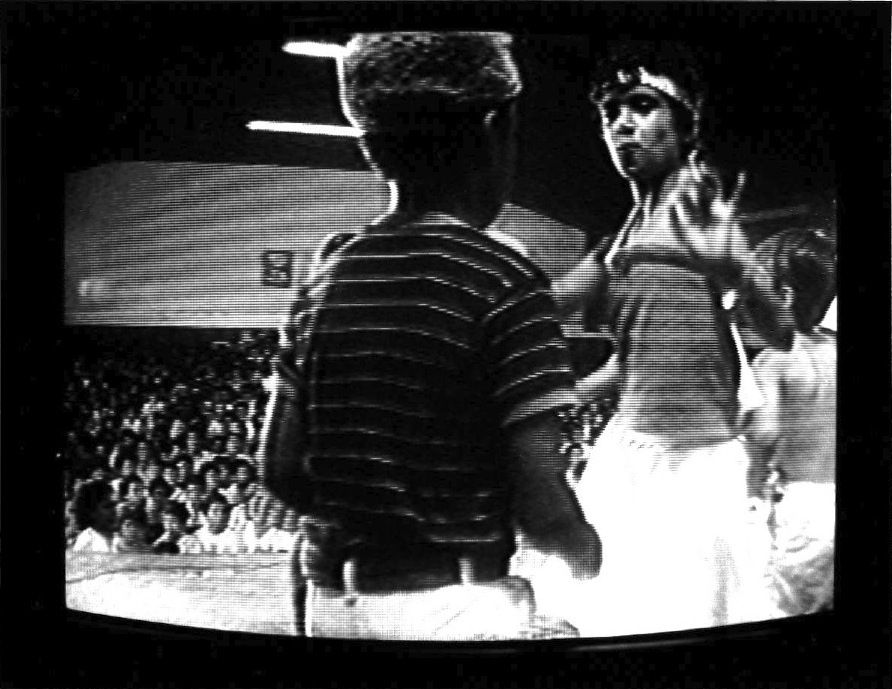 The back of one figured centered in the image as another figure appears in front of him alongisde a young shirtless child to the right and a seated audience is seen in the background. Scan lines in the image and the outer box of a CRT monitor are visible.