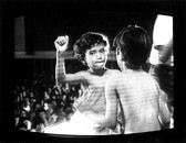 Two shirtless boys stand in front of each other, one with their back toward the viewer and the other facing the viewer. A partial view of taller figure is in the right side of the image and a crowd is seen seated in the background. Scan lines in the image and the outer box of a CRT monitor are visible.