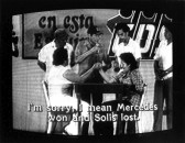 Two figures are sitting at the table with one hand tightly gripped to the other. Four men stand behind them looking on and partial text on the wall is visible in the background. Subtiltes burned onto the bottom of the image read,