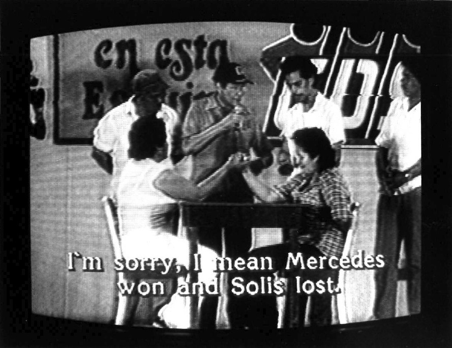 Two figures are sitting at the table with one hand tightly gripped to the other. Four men stand behind them looking on and partial text on the wall is visible in the background. Subtiltes burned onto the bottom of the image read,"I