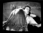 A man holds a woman over his shoulder as looks to the right. Writing can be seen on the wall behind them. Subtitles are burned into the bottom of the screen that read, 