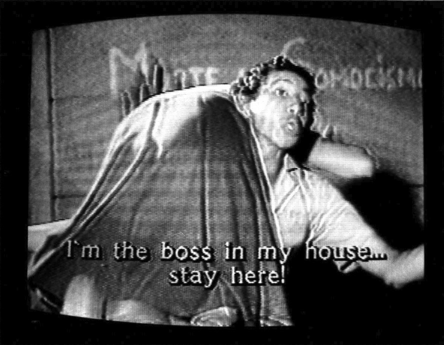 A man holds a woman over his shoulder as looks to the right. Writing can be seen on the wall behind them. Subtitles are burned into the bottom of the screen that read, "I