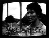 One figure on the left looks towards the camera and the other looks to the left. Outdoor suburban environment behind them. Subtitles burned into the bottom of the screen read, 