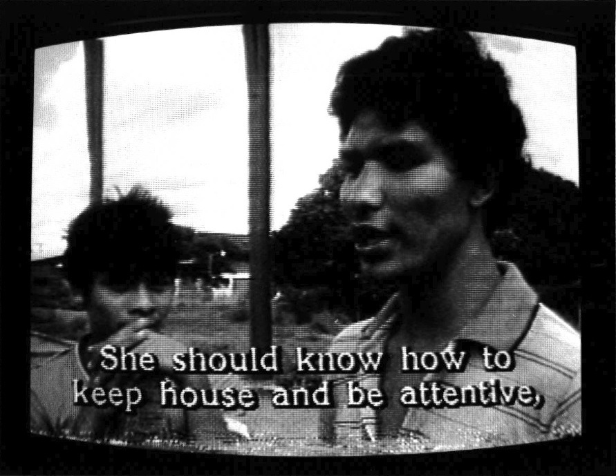 One figure on the left looks towards the camera and the other looks to the left. Outdoor suburban environment behind them. Subtitles burned into the bottom of the screen read, 