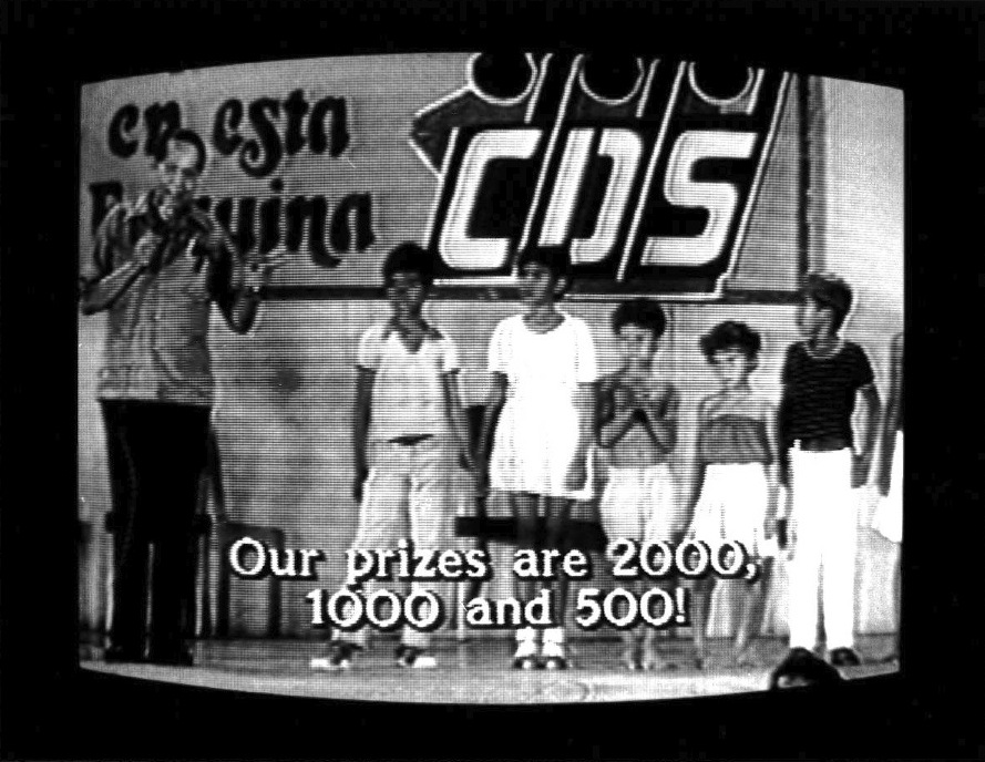 Older man with microphone stands next to five small boys. The wall behind them reads "en esta esquina" in black letters on one side and "CDS" in white letters on the other. Subtitles burned into the image at the bottom read, "Our prizes are 2000, 1000 and 500!" Video scan lines and outer edges of CRT monitor are visible.