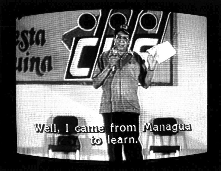 A figure standing central in the image with a microphone and two slightly upholstered chairs behind him. The logo "CDF" can be seen written on the wall next to another word of partial view. Video scan lines and black edges of CRT monitor are visible.