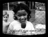 A small girl stands central in the image with a microphone near her mouth and a shirtless boy stands behind her to the left. A chain-link fence can be seen behind them next to a concrete building. Subtitles burned into the bottom of the image read, 