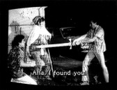 A figure holding a sword in the middle of a man and woman with a car visible behind them. Subtitles burned onto the bottom of the image read, 