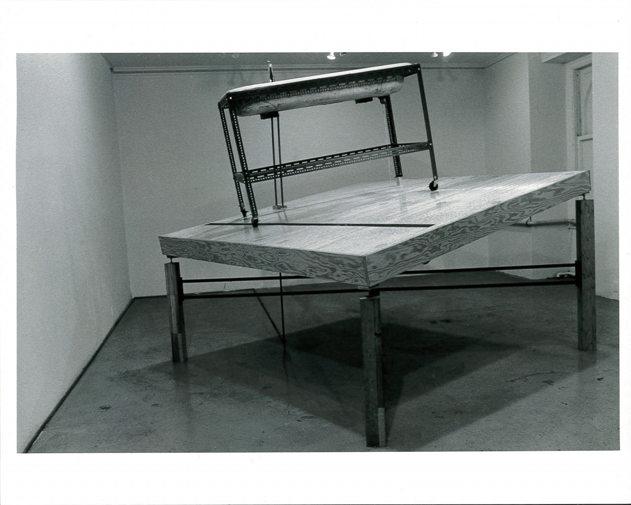 A sink structure on wheels sits atop a tilted wooden table top with legs of varying sizes. Light from the ceiling is seen emanating onto the structure in the white-walled gallery space.