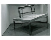 A sink structure on wheels sits atop a tilted wooden table top with legs of varying sizes. Light from the ceiling is seen emanating onto the structure in the white-walled gallery space.