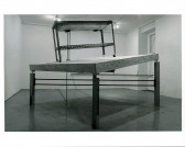 A sink structure on wheels sits atop a tilted wooden table top with legs of varying sizes. Light from the ceiling is seen emanating onto the structure in the white-walled gallery space.