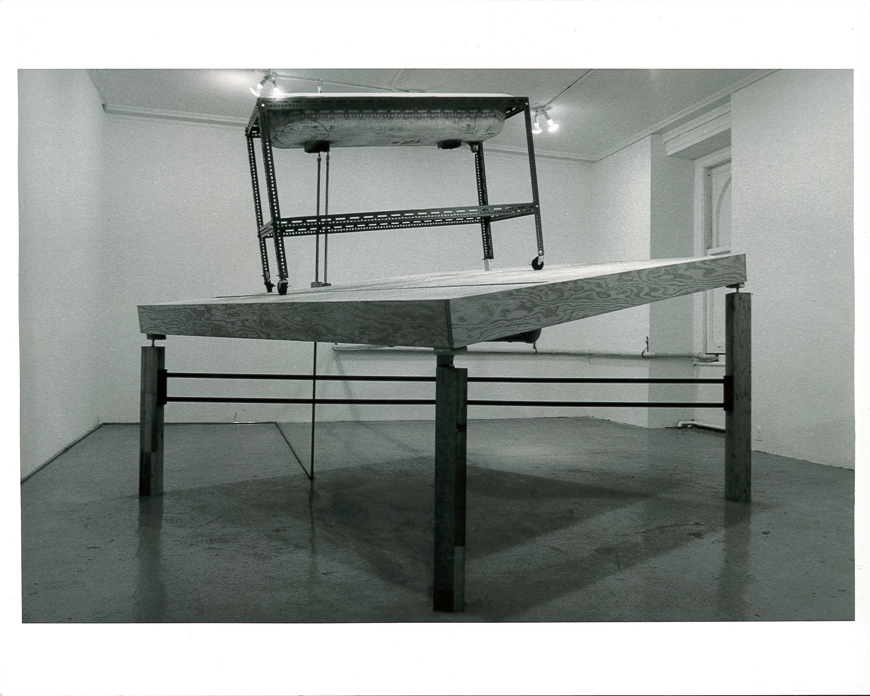 A sink structure on wheels sits atop a tilted wooden table top with legs of varying sizes. Light from the ceiling is seen emanating onto the structure in the white-walled gallery space.